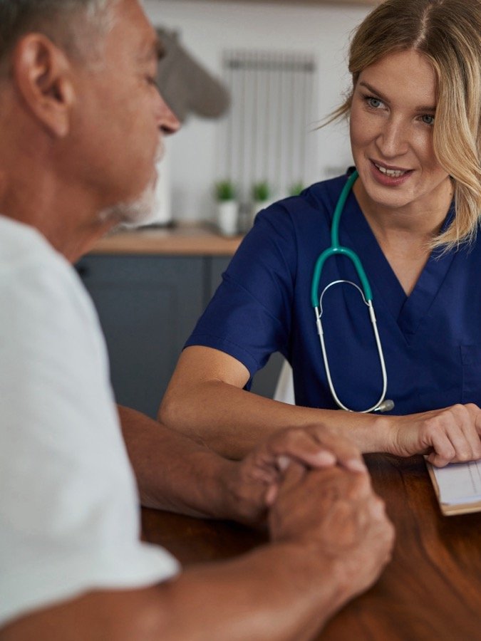 Médica conversando com paciente, segurando sua mão, à mesa da cozinha em casa